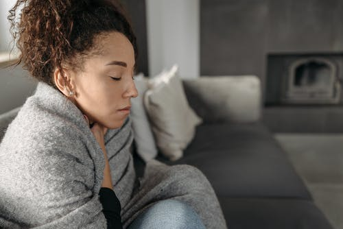 A woman who is sick sits on a couch, bundled up with her eyes closed.